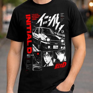 Pode incluir: Uma t-shirt preta de manga curta com uma impressão gráfica de um carro Toyota Sprinter Trueno AE86 branco, retratos de personagens em estilo manga japonês e texto incluindo INITIAL D, TOYOTA SPRINTER TRUENO AE86 e caracteres japoneses.