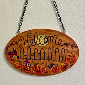 May include: Oval wooden Halloween welcome sign with a black chain for hanging. The sign features the word "Welcome" in cursive, a full moon, a picket fence, and several carved pumpkins with witch hats.