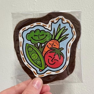 May include: A brown felt patch featuring a cheerful illustration of vegetables: broccoli, a carrot, a tomato, and peas. The patch is in a clear plastic sleeve, and the design is outlined with a stitched border.
