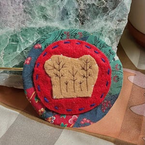 May include: A handmade fabric brooch with a forest design. The brooch features a red felt circle with a tan felt forest applique, stitched onto a patterned green and blue fabric base. The brooch is approximately 7.6 cm in diameter.