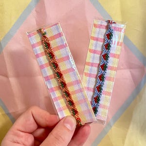May include: Two woven friendship bracelets featuring a repeating strawberry pattern. One bracelet has an orange background with green and red strawberries, while the other has a light blue background with green and red strawberries, each on a card.