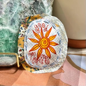May include: Handmade fabric patch with a sun design and the words "Good Morning." The patch features a yellow and orange sun on a white background with blue stitching. The patch is layered on patterned fabric.