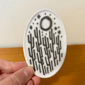 Peut inclure: Un écusson brodé de forme ovale avec un fond blanc et un motif graphique noir. Le design représente un paysage désertique stylisé avec plusieurs plantes verticales ressemblant à des cactus sous une lune circulaire et des étoiles.