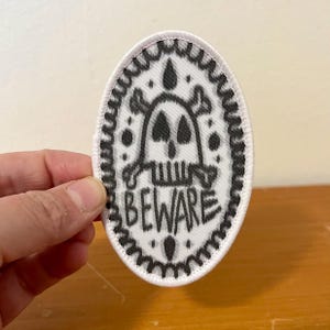 May include: An oval-shaped embroidered patch with a white background and a black border. The design features a central skull and crossbones graphic with the word BEWARE printed in black capital letters below it, surrounded by small black dots.