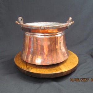 May include: A polished copper pot with a wide, rounded body and a metallic sheen. The vessel features two small, curved handles attached to the upper rim and a silver-toned interior lining. The pot has a smooth, reflective surface finish.