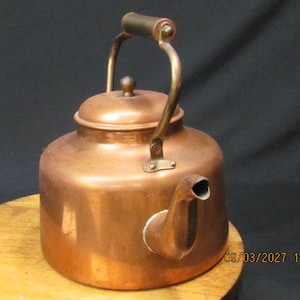 May include: A copper kettle with a rounded body, a spout, and a handle. The kettle has a lid with a small knob. The handle is attached to the body with brass fittings. The kettle is sitting on a wooden surface.