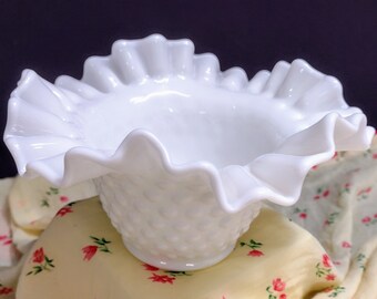 May include: A white milk glass bowl with a ruffled edge and a textured, dimpled surface. The bowl is sitting on a floral patterned fabric.