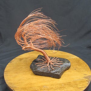 May include: A copper wire sculpture of a tree with flowing branches and roots, mounted on a dark gray stone. The tree's delicate form is crafted from thin copper wire, creating a unique artistic representation of nature.