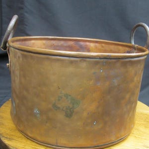 May include: A copper-colored, oval-shaped bucket with two arched handles. The bucket has a hammered texture and a slightly tarnished appearance, suggesting age. The handles are made of a lighter-colored metal.
