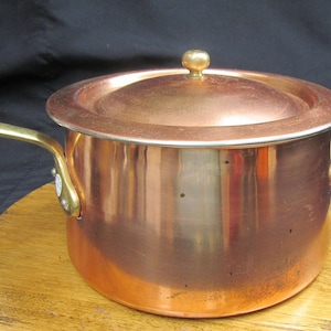May include: A copper saucepan with a lid and a brass handle. The pot has a shiny, reflective surface and a rounded shape. The lid has a small brass knob. The saucepan is sitting on a wooden surface.