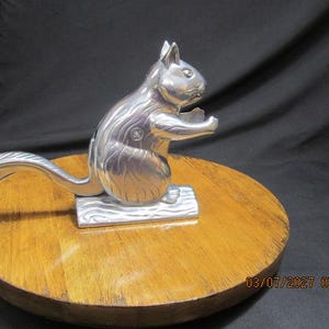 May include: A silver-colored metal nutcracker shaped like a squirrel, featuring a long curved tail that acts as a lever. The squirrel is depicted in a seated position on a textured base, with its front paws raised. The date 03/07/2027 is visible.