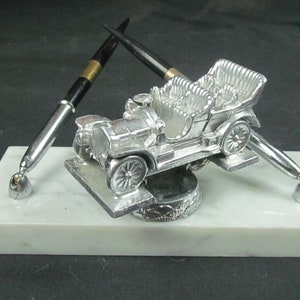 May include: A silver vintage car model with two black and silver pens on a white marble base.