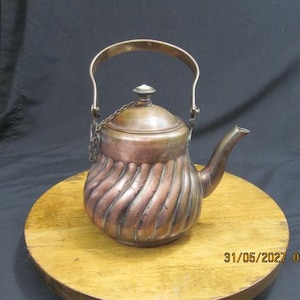 May include: A copper kettle with a rounded, spiraled body, a curved spout, and a hinged lid featuring a small white knob. The kettle includes a tall, arched metal handle attached to the sides, with a thin metal chain connecting the lid to the handle.