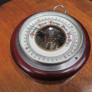 May include: A round analog barometer with a dark polished wood frame and a white dial. The dial features black numerical markings and the text Stormy, Rain, Change, Fair, and Very Dry in red and black lettering. A metal needle points to the left.