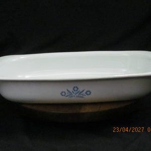 May include: White rectangular baking dish with rounded corners and a blue floral design. The dish is made of ceramic and has a slightly raised rim. The dish is sitting on a wooden surface.