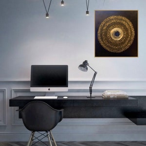 May include: A black and gold abstract painting with a circular design. The painting is hanging on a white wall in a home office setting. A black desk with a computer, lamp, and books is in the foreground.
