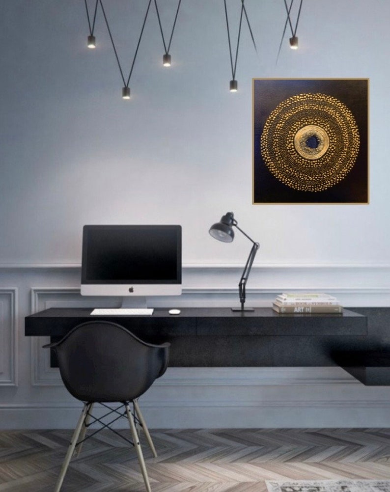 May include: A black and gold abstract painting with a circular design. The painting is hanging on a white wall in a home office setting. A black desk with a computer, lamp, and books is in the foreground.