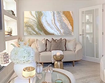 May include: A white couch with gold accents sits in a living room with a large abstract painting featuring white, gold, and silver colors. A white vase with white flowers sits on a glass coffee table with a gold trim.