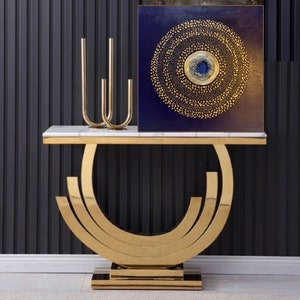 May include: A gold metal console table with a white marble top. The table has a unique curved design and features a gold metal base with three tiers. A gold metal candle holder with three arms is on the table, along with a square canvas painting with a blue background and a gold circle design.