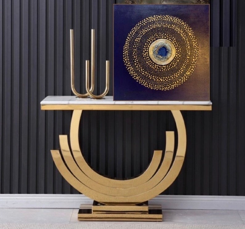 May include: A gold metal console table with a white marble top. The table has a unique curved design and features a gold metal base with three tiers. A gold metal candle holder with three arms is on the table, along with a square canvas painting with a blue background and a gold circle design.