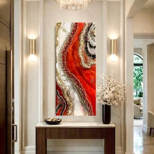May include: Abstract resin art piece with a vibrant red, gold, black, and white design. The artwork is displayed on a wall in a well-lit hallway, above a wooden console table. The room features a chandelier and sconces.