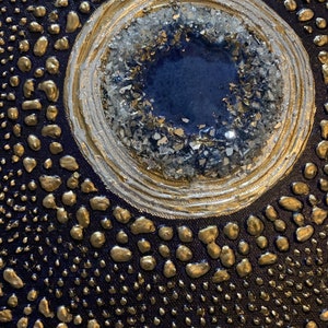 May include: A close-up of a blue and gold abstract painting with a circular design. The center of the circle is filled with a blue resin and surrounded by gold and white paint. The background is a dark blue canvas with gold paint splatters.