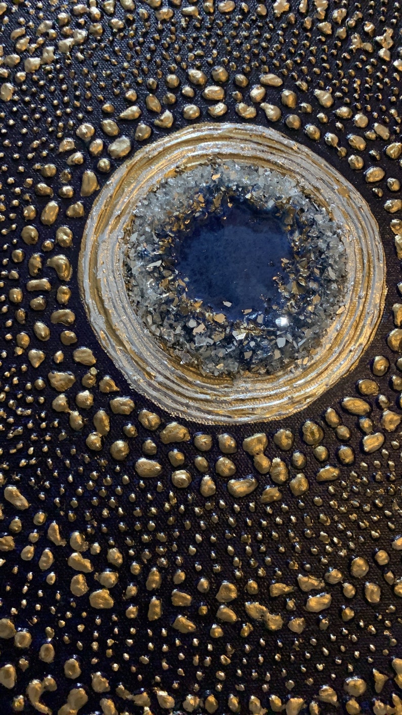 May include: A close-up of a blue and gold abstract painting with a circular design. The center of the circle is filled with a blue resin and surrounded by gold and white paint. The background is a dark blue canvas with gold paint splatters.