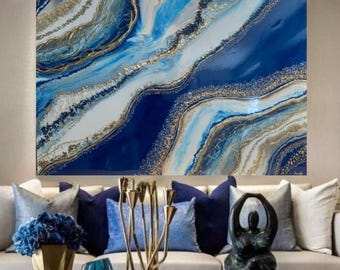May include: Abstract painting with blue, white, and gold swirls and glitter. The painting is framed and hanging on a wall above a couch with blue pillows.