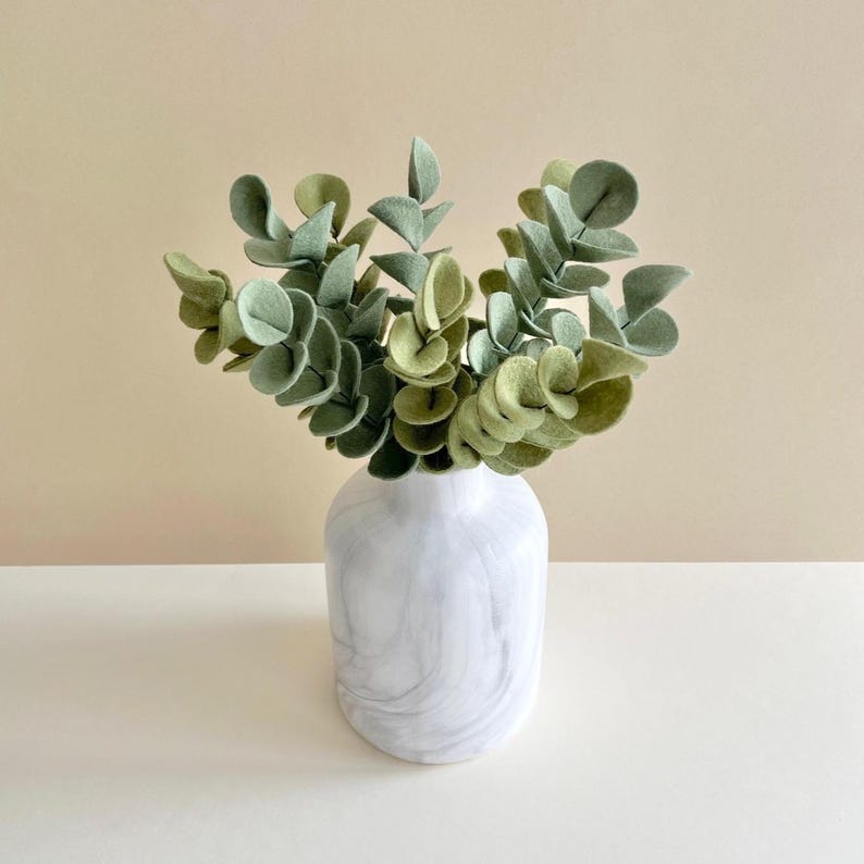 May include: A white marble-patterned vase containing a cluster of artificial eucalyptus stems with rounded, felt-like leaves in various shades of sage green and olive green, arranged in a dense, upright bouquet with a smooth, rounded base.