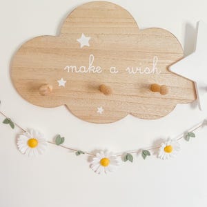 Puede incluir: Decoraci&oacute;n de pared de madera con forma de nube con el texto "make a wish" y tres clavijas de madera. La nube tiene detalles de estrellas blancas. Una guirnalda de margaritas con flores blancas y centros amarillos cuelga debajo.