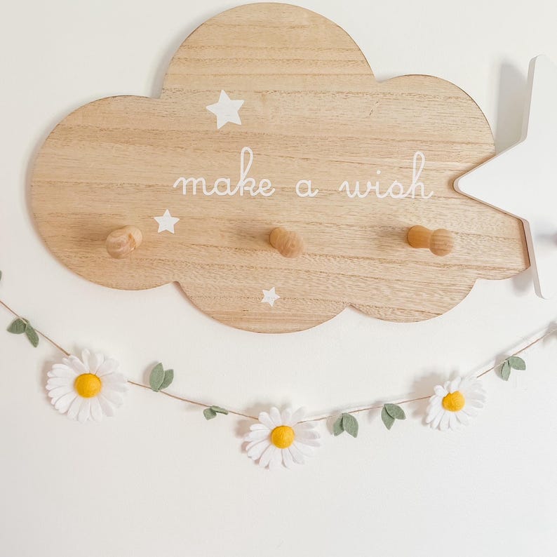 Puede incluir: Decoraci&oacute;n de pared de madera con forma de nube con el texto "make a wish" y tres clavijas de madera. La nube tiene detalles de estrellas blancas. Una guirnalda de margaritas con flores blancas y centros amarillos cuelga debajo.