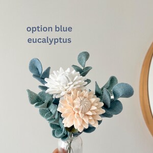 Idée cadeau fait main pour mère bouquet de fleurs en feutrine tendance décoration d'intérieur plante en laine  fleur en tissu pour la maison Cadeau Fête des Mères bouquet de fleurs en feutrine cadeau d'anniversaire pour maman ou épouse déco mason en laine idée bouquet de printemps Un petit bouquet de fleurs en feutre et de feuilles d'eucalyptus dans un vase en verre transparent. Les fleurs sont blanches et pêche, avec des feuilles d'eucalyptus bleu-vert.