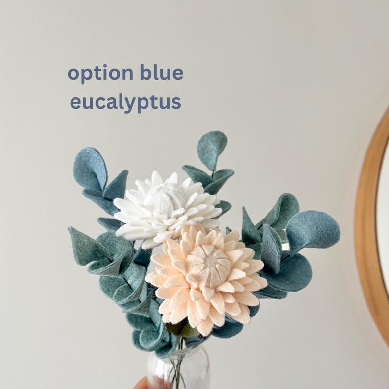 Idée cadeau fait main pour mère bouquet de fleurs en feutrine tendance décoration d'intérieur plante en laine  fleur en tissu pour la maison Cadeau Fête des Mères bouquet de fleurs en feutrine cadeau d'anniversaire pour maman ou épouse déco mason en laine idée bouquet de printemps Un petit bouquet de fleurs en feutre et de feuilles d'eucalyptus dans un vase en verre transparent. Les fleurs sont blanches et pêche, avec des feuilles d'eucalyptus bleu-vert.