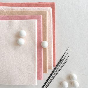 Pode incluir: Uma pilha de folhas de feltro retangulares em tons de rosa claro, p&ecirc;ssego e coral, acompanhada por v&aacute;rias pequenas bolas de feltro brancas e um grupo de agulhas pretas finas. As folhas de feltro est&atilde;o sobrepostas em um gradiente de cores.