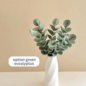 Op de afbeelding: Een witte gedraaide vaas bevat verschillende stelen van vilt eucalyptusbladeren in een zachte saliegroene kleur. Een wit rechthoekig label aan de linkerkant van de afbeelding bevat de donkerblauwe tekst die option green eucalyptus luidt.