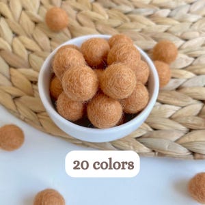 May include: A white ceramic bowl filled with multiple light brown felt balls, with additional loose felt balls scattered nearby. A white rectangular label with rounded corners displays the text 20 colors in brown font.