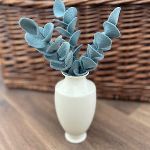 May include: A small, smooth, off-white ceramic vase containing several stems of artificial blue-green felt eucalyptus leaves. The vase has a narrow neck and a flared base, holding the fabric foliage in an upright, branching arrangement.