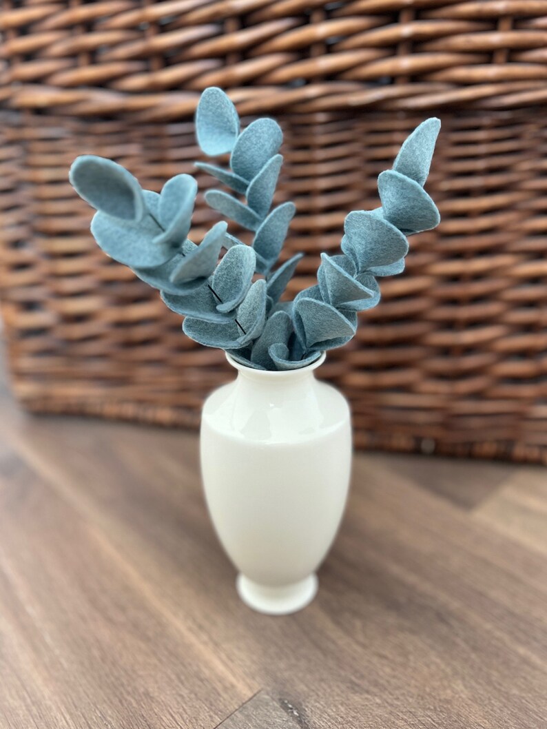 May include: A small, smooth, off-white ceramic vase containing several stems of artificial blue-green felt eucalyptus leaves. The vase has a narrow neck and a flared base, holding the fabric foliage in an upright, branching arrangement.