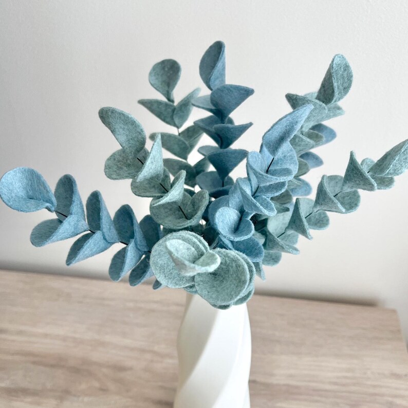 May include: A decorative arrangement of multiple stems of felt eucalyptus leaves in a soft, muted blue-green color. The leaves are rounded and arranged in a symmetrical pattern along each stem, creating a full, textured botanical display.
