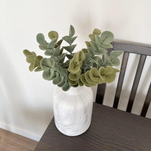 May include: A white marble-patterned vase containing several stems of felt eucalyptus leaves in varying shades of sage and olive green. The plant stems are arranged upright, showcasing rounded, soft-textured foliage in a dense, layered cluster.
