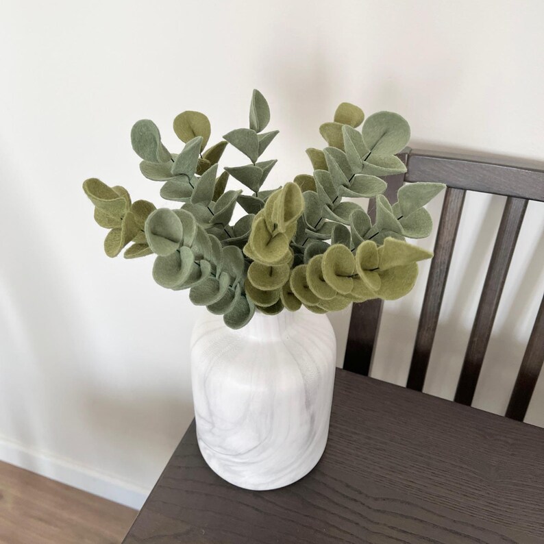 May include: A white marble-patterned vase containing several stems of felt eucalyptus leaves in varying shades of sage and olive green. The plant stems are arranged upright, showcasing rounded, soft-textured foliage in a dense, layered cluster.