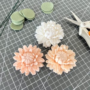 Op de afbeelding: Drie handgemaakte viltbloemen in wit en perzik, vergezeld door verschillende kleine saliegroene viltcirkels, dunne groene draadstelen en een schaar met witte handgrepen en een metalen blad op een donkergrijze rastermat.