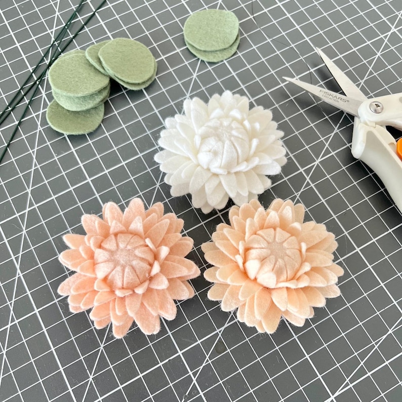 Op de afbeelding: Drie handgemaakte viltbloemen in wit en perzik, vergezeld door verschillende kleine saliegroene viltcirkels, dunne groene draadstelen en een schaar met witte handgrepen en een metalen blad op een donkergrijze rastermat.