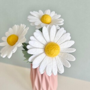 May include: Three felt daisy flowers with bright yellow centers and white petals arranged in a light pink vase with a vertical ribbed texture. The flowers feature soft, rounded textures and are positioned to show their full circular blooms.
