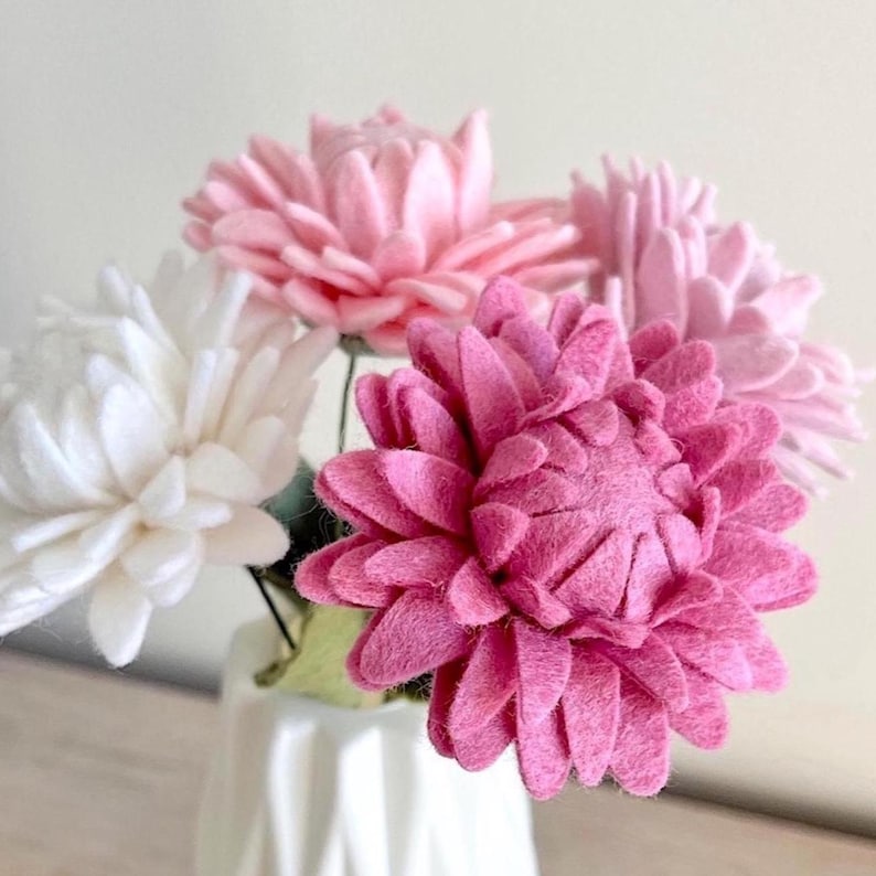 May include: A bouquet of felt flowers in shades of white, light pink, and dark pink. Each flower features layered, rounded petals arranged in a dense, circular pattern. The flowers are supported by thin green stems and are held in a white vase.