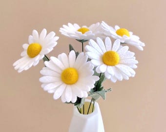May include: A bouquet of five artificial daisy flowers with white felt petals and bright yellow centers. Each flower is attached to a thin green stem with small green leaves, and the entire arrangement is displayed in a white vase.