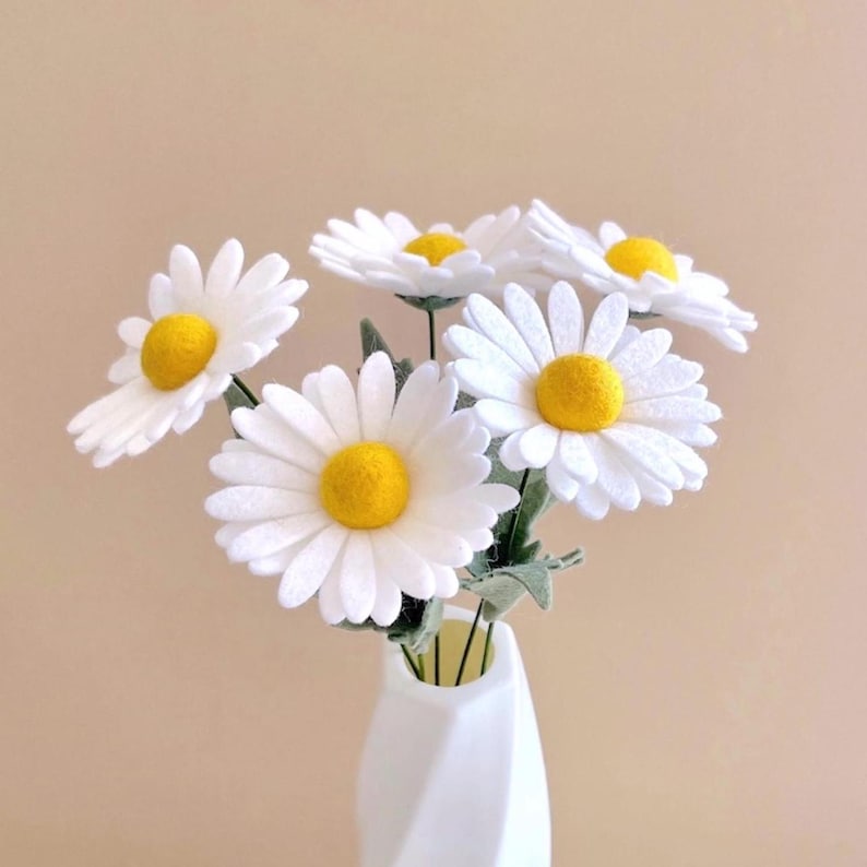 May include: A bouquet of five artificial daisy flowers with white felt petals and bright yellow centers. Each flower is attached to a thin green stem with small green leaves, and the entire arrangement is displayed in a white vase.