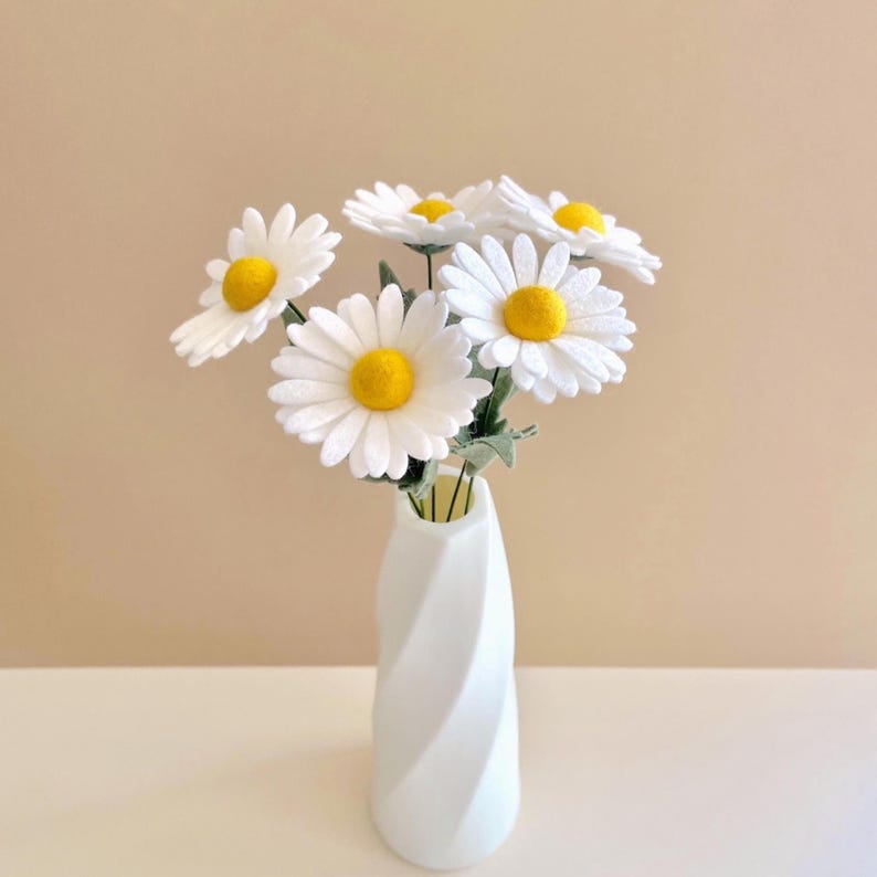May include: A bouquet of five artificial daisy flowers with white petals and bright yellow centers, arranged in a white ceramic vase featuring a spiraled, twisted design. The stems are thin and green, supporting the flower heads.