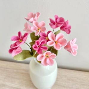 Op de afbeelding: Een boeket van handgemaakte viltbloemen in diverse roze tinten, variërend van licht blush tot diep magenta, elk met een klein geel hartje. De bloemen zijn bevestigd aan dunne groene stengels met vilten bladeren en staan in een witte vaas.