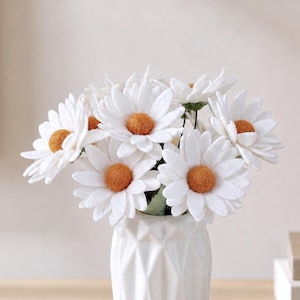 Op de afbeelding: Een boeket kunstmatige madeliefjes met witte viltblaadjes en gestructureerde goudgele harten. De bloemen staan in een witte keramische vaas met een geometrisch, gefacetteerd oppervlakontwerp met hoekige, diamantvormen.